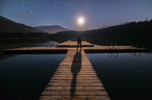 明るい月夜に、湖のほとりにたたずむ男性の写真